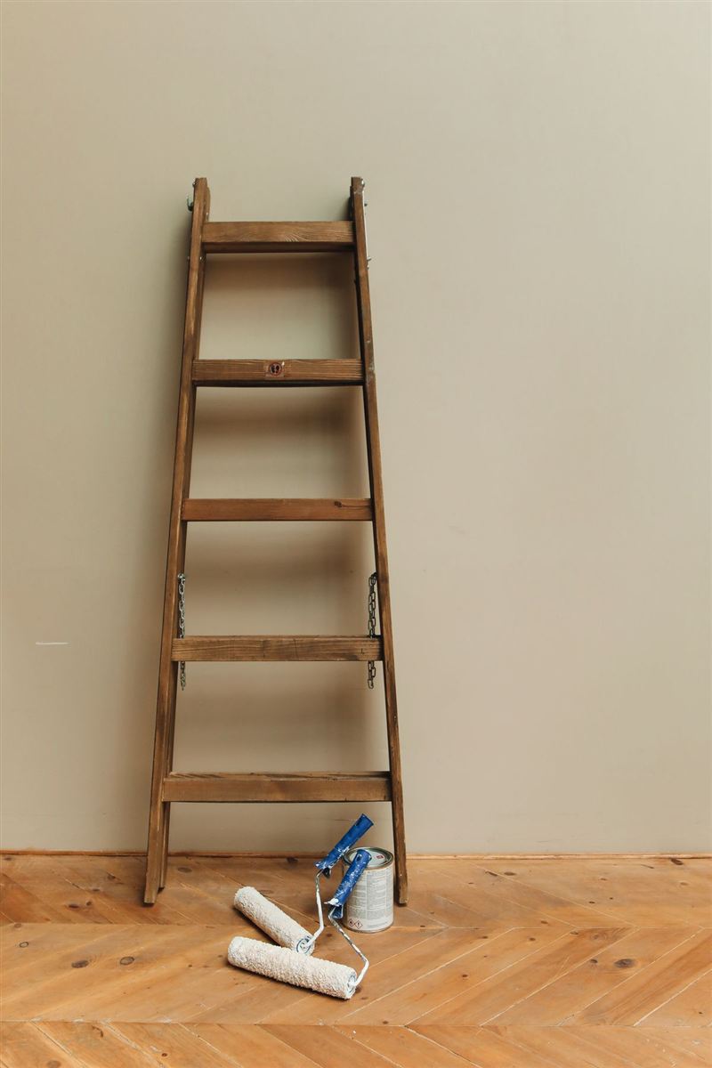 A rustic wooden ladder leaning against a beige wall, with two cream paint rollers and a paint can on a herringbone wood floor.